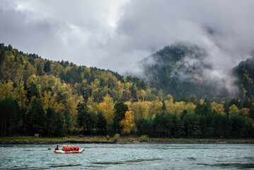 An inflatable rubber boat with rafters in red life jackets and yellow helmets are rafting on a mountain river, against the background of bright autumn mountains covered with thick fog and clouds