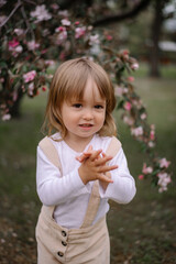 A small stylish girl, a child in beige trousers and a white blouse, claps her hands and looks into the distance. Against the background of a blooming apple tree in a green garden