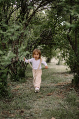 A small stylish girl, a child in beige trousers and a white blouse, runs in a tunnel from tall green bushes