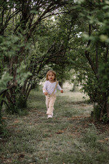 A small stylish girl, a child in beige trousers and a white blouse, runs in a tunnel from tall green bushes