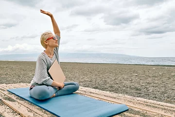 Fototapeten Positive and fit blonde woman in sportswear, sitting in lotus pose, with laptop at the beach waiting for friends. Freelance work, wellness concept. © Caterina Trimarchi