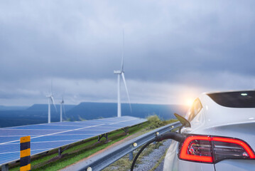Electric car plugged into a charging station with wind turbines and solar panels in the background,...