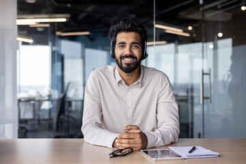 Portrait of a smiling Muslim man businessman, marketer, salesman sitting in the office at the table in a headset and smiling confidently at the camera - Powered by Adobe