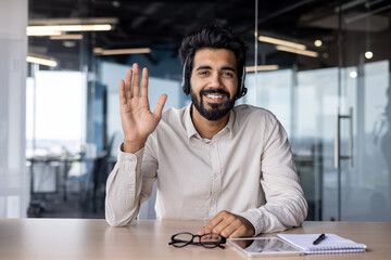 Indian young smiling businessman sitting in the office at the desk in front of the camera in the headset, waving and greeting with his hand to the camera. Close-up portrait