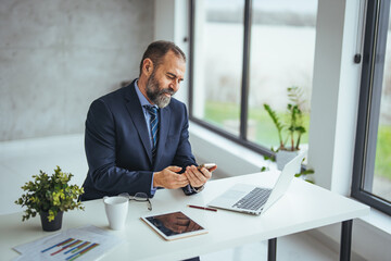 Mature business man in formal clothing wearing spectacles using mobile phone. Serious businessman using smartphone and digital tablet at work. Manager in suit using cellphone in a modern office.