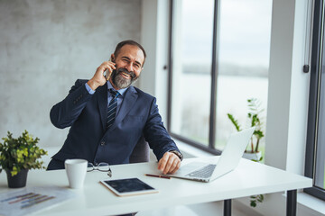 Smiling businessman wearing glasses talking on phone, sitting at desk with laptop, friendly manager...