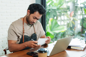 Indian man and businessman with beard Barista, coffee shop owner Sit and work in front of a tablet,...