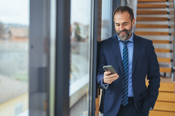 Handsome businessman wearing eyeglasses using smart phone and smiling while standing near the office window, Copy Space. Handsome businessman dressed in the suit reading with his smart phone