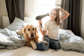 Cute Little Girl Drying Hair With A Hairdryer, Sitting With Dog On A Bed