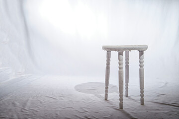 Solitary Wooden Stool in a White Minimalist Setting. Lone, white-painted wooden stool stands centered on a textured surface, surrounded by soft white backdrop - obrazy, fototapety, plakaty