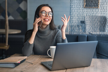 Happy business woman talking on cell phone sitting at table and working. Asian female sitting at office having telephonic conversation with client and smiling with look away