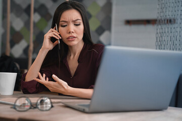 Busy woman sitting at her workplace and discussing with gesticulation some job questions on mobile phone in hand. Multitasking, deadline and workload