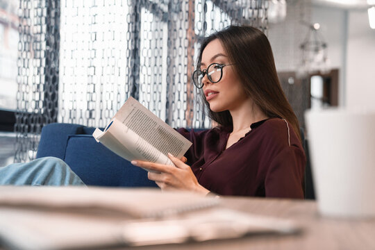 Attractive woman in eyeglasses reading book at café with cup and documents on foreground. Break after work, preparing for exams, doing homework, e-learning, studying remotely