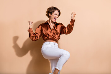 Photo of lucky impressed woman wear brown blouse rising fists screaming eyes empty space isolated beige color background
