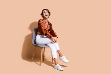 Full length photo of cute excited woman wear brown blouse sitting chair looking up empty space isolated beige color background