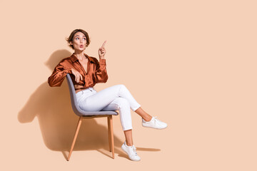 Full length photo of excited impressed woman wear brown blouse sitting chair looking pointing empty space isolated beige color background