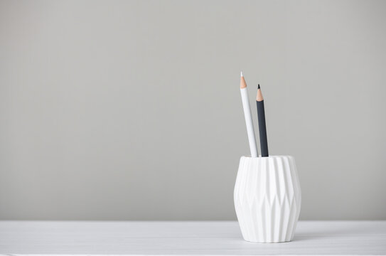 Two white and gray pencils stand in a glass on a white table on a gray background in a minimalist style, space for text, concept of office, learning, papers, development, growth, stationery