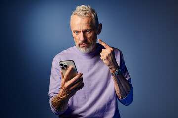 handsome bearded fashionable mature man in purple stylish sweatshirt grimacing and holding phone