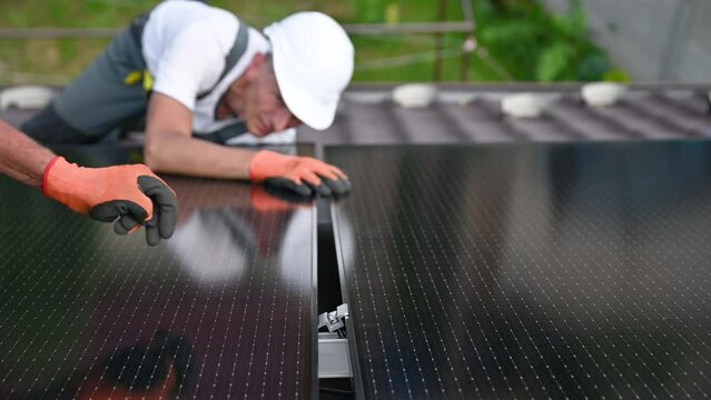 Workers building solar panel system on rooftop of house. Two men installers in gloves installing photovoltaic solar module outdoors. Alternative, green and renewable energy generation concept.