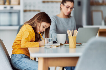 A cute girl sits at the dining table with her busy mother and draws.