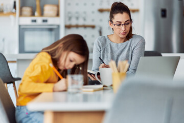 A serious woman working from home on a laptop while her daughter draws.