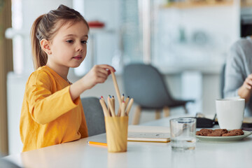 A cute girl sits at the table with cookies and draws with wooden crayons.