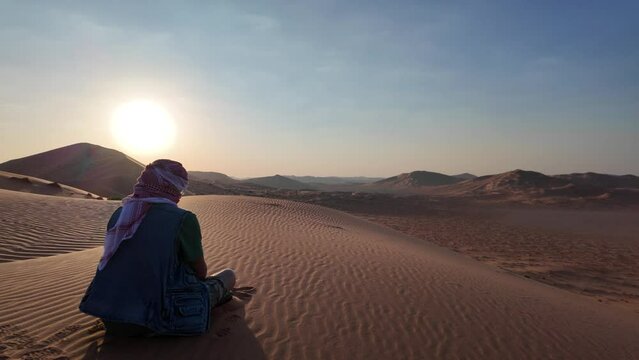 beduin man in vastness of the Empty Quarter with kefiah hat in contrast to the golden sand at dawn. He seeks adventure and solitude in land of Oman Rub' al-Khali desert.