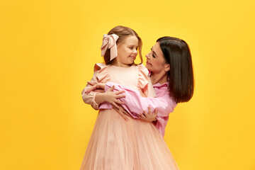 Happy, smiling, beautiful young woman hugging her adorable child, daughter against yellow studio background. Concept of happiness, Mother's day, childhood, fashion and lifestyle