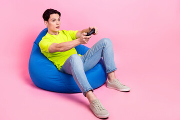 Full length photo of cool impressed man dressed yellow t-shirt sitting bean bag enjoying playstation game empty space isolated pink color background - obrazy, fototapety, plakaty
