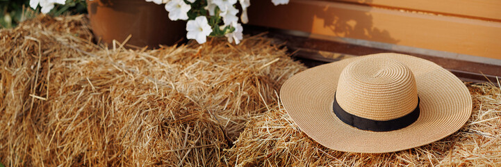 Country scene with straw hat on hay bales, blooming white flowers and wooden background. Country living or farmhouse style decor concept. Banner with copy space.