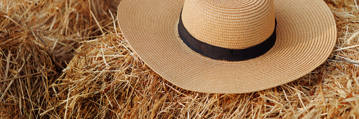 A straw hat with a black ribbon rests on a pile of hay in a rustic outdoor setting. Concept for fashion, rural life, or summer vibes. Plenty of copy space. Banner with copy space.