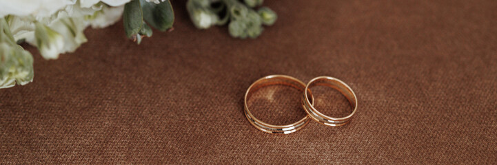 Golden wedding rings lay near a lush, white bouquet on a textured brown surface. Concept for wedding invitations or bridal shops. Banner with copy space.