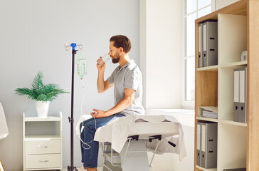 Young man sitting on the couch at doctor's office in clinic drinking a glass of water while...