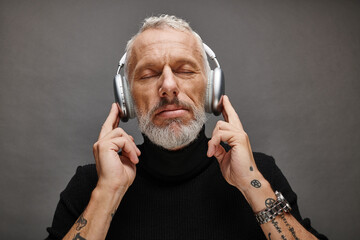 joyful appealing mature man in voguish turtleneck with gray beard and headphones sitting on chair