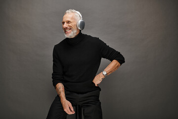 joyful appealing mature man in voguish turtleneck with gray beard and headphones sitting on chair