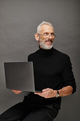 joyous attractive bearded mature man with glasses sitting on chair with laptop and smiling happily - 731004954