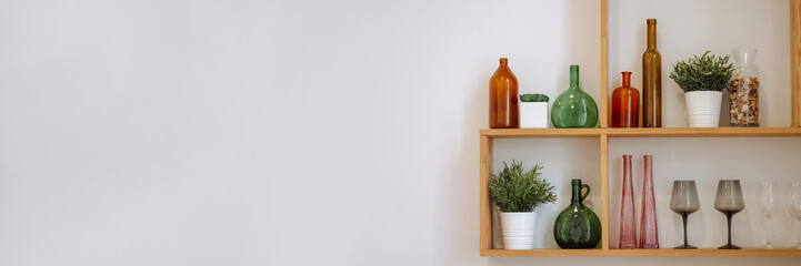 Minimalist wooden shelves displaying amber and green bottles, clear glasses, and potted greenery...