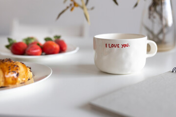 White cup with the inscription I love you, croissant and strawberries on a white breakfast table. Bouquet with dried mimosa.