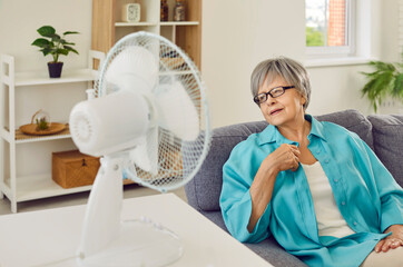 Senior woman trying to escape from summer heat by electric fan at home. Mature lady sitting on sofa...