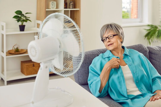 Senior woman trying to escape from summer heat by electric fan at home. Mature lady sitting on sofa in living room, sweating, breathing hard, and trying to refresh with blowing electric ventilator