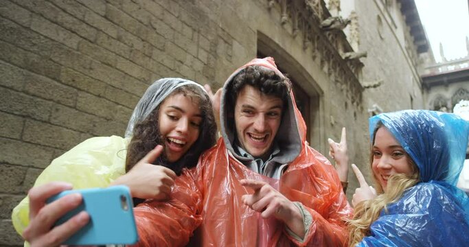 Friends, people and selfie in street, rain and funny face with smile, thumbs up and excited for live stream. Group, gen z women and man with photography, choice or post on social media in Barcelona