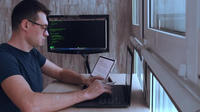 Young handsome male programmer in glasses sits at a laptop typing code looking at a tablet. Freelancing concept.
