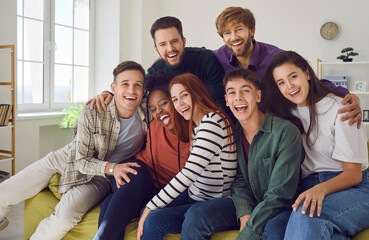 Fototapeta premium Group of young people sits on a sofa at home together, sharing moments of laughter and happiness. Portrait of a diverse camaraderie of friends, smiling and looking into the camera.