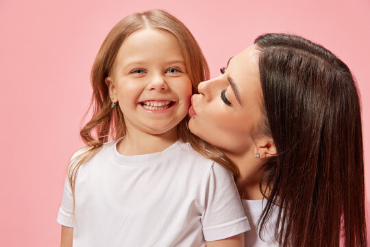 Caring, happy young woman, mother kissing her cute, smiling, tender little girl, daughter against pink studio background. Concept of happiness, Mother's day, childhood, fashion and lifestyle