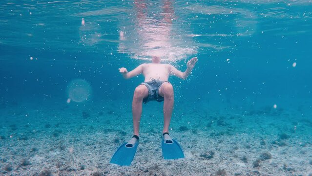 A caucasian tall guy in snorkeling mask underwater grabs flippers with his hands against background of beautiful reef bottom. A young man on vacation fooling around and having fun at bottom of ocean