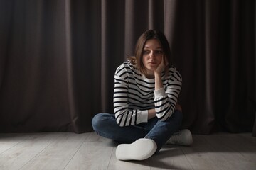 Sad young woman sitting on floor near curtains