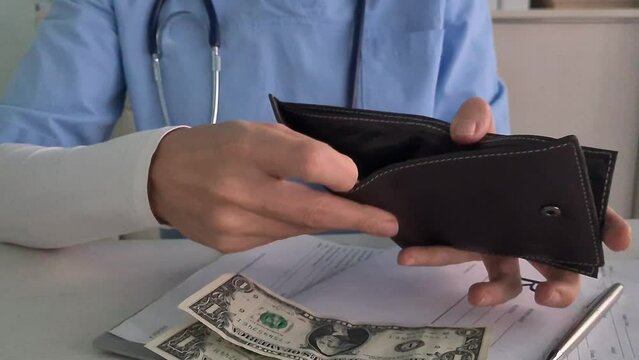 Doctor wearing a white coat is holding an empty wallet, and there is a prescription pad and a pen on the table