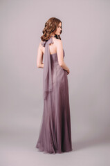 Elegant purple sleeveless evening dress with back bow, rear view. Beautiful lilac maxi chiffon gown. Studio sensual portrait of a young brunette woman. Summer shoulder off female look for an event.