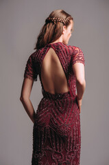 Fototapeta premium Bead embellished floral short sleeve maxi dress with back cut in maroon. Red backless transparent evening gown with a train. Beautiful young brunette woman's studio portrait. Female elegant clothes.