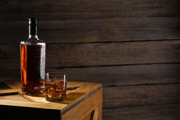 Whiskey with ice cubes in glass and bottle on wooden crate, space for text